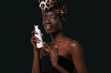 Smiling black girl holding bottle with cosmetic cream. Beautiful young woman wearing traditional african turban and tank top. Body and skin care. Isolated on black background. Studio shoot. Copy space