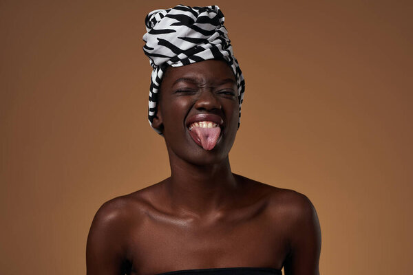 Joking elegant black girl wearing traditional african turban winking and sticking out tongue. Beautiful young slim woman. Female beauty. Isolated on orange background. Studio shoot. Copy space