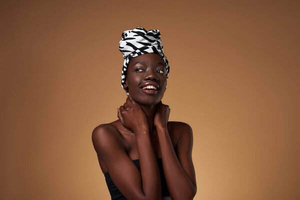 Smiling fashionable black girl wearing traditional african turban looking away. Pretty young slim woman wearing tank top. Female beauty. Isolated on orange background. Studio shoot. Copy space
