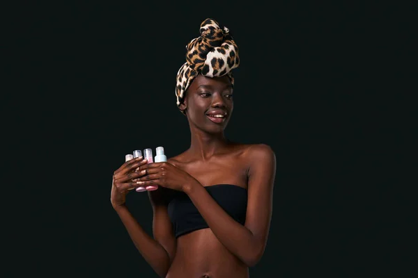 Smiling elegant black girl holding four colorful nail polishes. Beautiful young woman wearing traditional african turban and tank top. Body and skin care. Black background. Studio shoot. Copy space