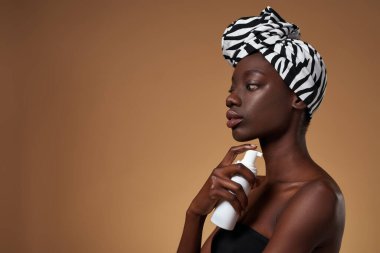 Üzerinde kozmetik krem olan bir şişe tutan düşünceli zenci kız. Geleneksel Afrika sarığı ve kolsuz bluz giyen zayıf bir kadın. Vücut ve cilt bakımı. Turuncu arka plan. Stüdyo çekimi. Boşluğu kopyala