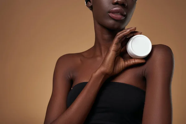 Obscure face of fashionable black girl holding container with cosmetic cream. Pretty young slim woman wearing and tank top. Body and skin care. Isolated on orange background. Studio shoot. Copy space