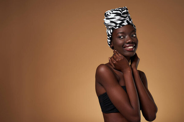 Smiling elegant black girl wearing traditional african turban looking away. Beautiful young slim woman wearing tank top. Female beauty. Isolated on orange background. Studio shoot. Copy space