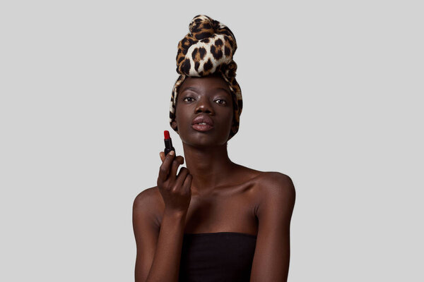 Elegant black girl holding lipstick and looking at camera. Beautiful young slim woman wearing traditional african turban and tank top. Female beauty. White background. Studio shoot. Copy space