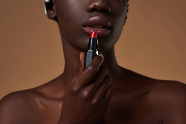 Cropped of african american girl painting lips with red lipstick. Obscure face of attractive young slim woman. Female beauty. Isolated on orange background. Studio shoot