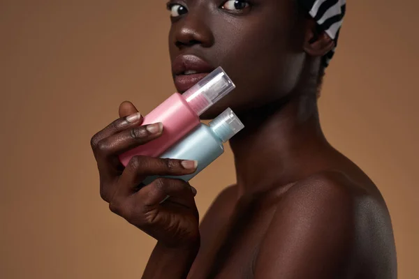 Partial side view of elegant black girl holding colorful nail polishes and looking at camera. Obscure face of youngwoman wear traditional african turban. Orange background. Studio shoot. Copy space