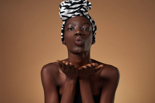 Stylish black girl wearing traditional african turban blowing air kiss and looking at camera. Attractive young slim woman wearing tank top. Female beauty. Orange background. Studio shoot. Copy space