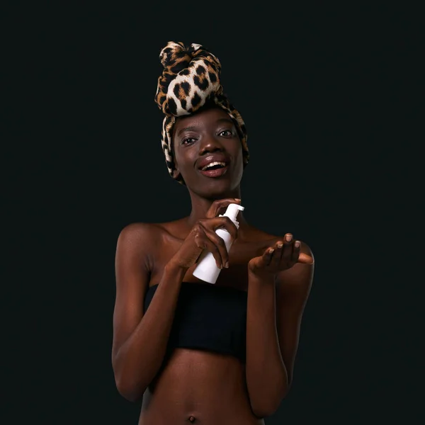 Black girl pouring cosmetic cream on palm hand and looking at camera. Young slim woman wearing traditional african turban and tank top. Body and skin care. Black background. Studio shoot. Copy space