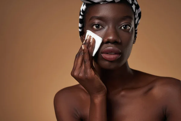 Partial of black girl wiping face with cotton swab and looking at camera. Young woman wearing traditional african turban. Hygiene and skin care. Orange background. Studio shoot. Copy space