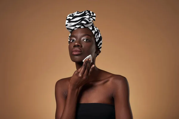 Black girl wiping her face with cotton swab and looking at camera. Young slim woman wearing traditional african turban and tank top. Hygiene and skin care. Orange background. Studio shoot. Copy space