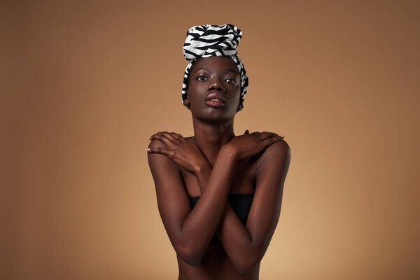 Front view of serious black girl wearing traditional african turban hugging herself. Pretty young slim woman wearing tank top. Female beauty. Isolated on orange background. Studio shoot. Copy space