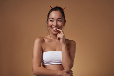 Kurnaz Asyalı kız parmağını ısırıyor ve başka tarafa bakıyor. Güzel, genç, esmer, zayıf, kolsuz bluz giyen kadın. Kadın güzelliği. Turuncu arka planda izole edilmiş. Stüdyo çekimi. Boşluğu kopyala