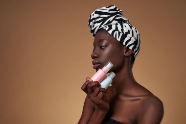 Stylish black girl holding colorful nail polishes. Attractive young slim woman wearing traditional african turban and tank top. Female beauty. Isolated on orange background. Studio shoot. Copy space