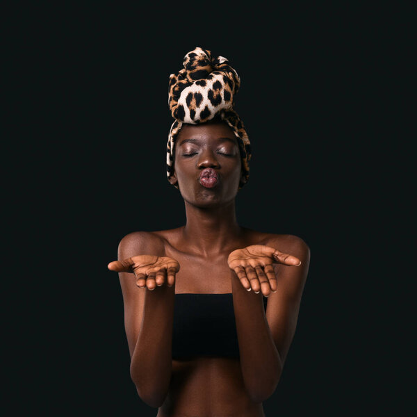 Front view of black girl wearing traditional african turban kissing with outstretched hands. Pretty young slim woman with closed eyes. Female beauty. Isolated on black background in studio. Copy space