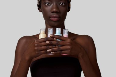Cropped of serious stylish black girl holding four colorful nail polishes. Young slim woman wearing traditional african turban and tank top. Female beauty. White background. Studio shoot. Copy space