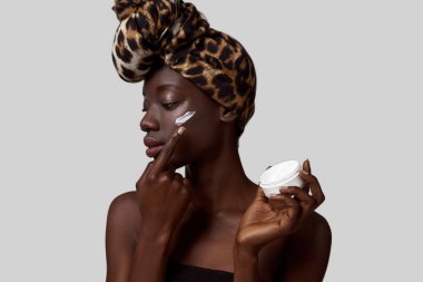 Elegant black girl smearing cosmetic cream from container on her face. Beautiful young slim woman wearing traditional african turban. Body and skin care. White background. Studio shoot. Copy space