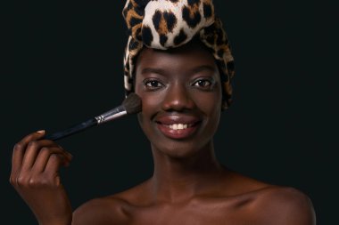 Portrait of smiling stylish black girl with cosmetic brush. Attractive young slim woman wearing traditional african turban. Female beauty. Isolated on black background. Studio shoot. Copy space