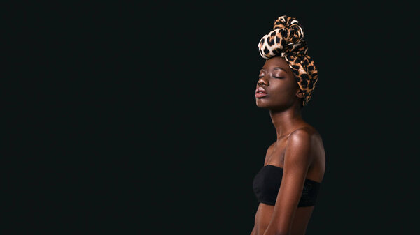 Side view of dreamy stylish black girl wearing traditional african turban. Attractive young slim woman wearing tank top with closed eyes. Female beauty. Dark background. Studio shoot. Copy space