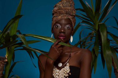 Fashionable black girl between green palms looking at camera. Pretty young slim woman wearing tank top and traditional african turban, necklace and earrings. Isolated on blue background. Studio shoot