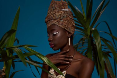 Thoughtful stylish black girl posing between green palms. Attractive young slim woman wearing tank top and traditional african turban, necklace and earrings. Isolated on blue background. Studio shoot