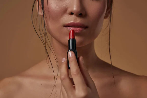 Obscure face of girl holding red lipstick. Partial of attractive young brunette woman. Concept of female beauty. Isolated on orange background. Studio shoot