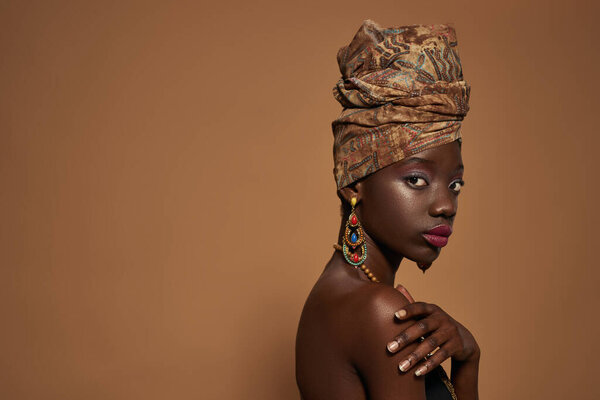 Side view of serious elegant black girl wearing traditional african outfit and accessories looking at camera. Cropped of beautiful young slim woman. Isolated on orange background in studio. Copy space