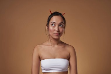 Front view of elegant asian girl thinking about something. Beautiful young brunette slim woman wearing tank top. Female beauty. Isolated on orange background. Studio shoot. Copy space
