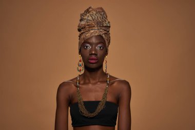 Front view of serious elegant black girl wearing traditional african outfit and accessories looking at camera. Beautiful young slim woman. Isolated on orange background. Studio shoot. Copy space