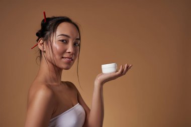 Side view of elegant asian girl holding container with cosmetic cream on hand palm. Beautiful young brunette slim woman. Body and skin care. Isolated on orange background. Studio shoot. Copy space