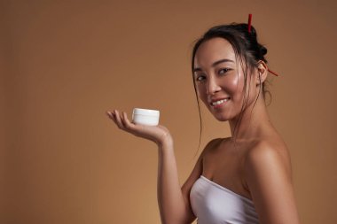 Side view of smiling asian girl holding container with cosmetic cream on hand palm. Attractive young brunette woman looking at camera. Body and skin care. Orange background. Studio shoot. Copy space