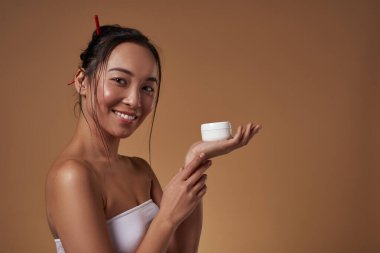 Smiling asian girl holding container with cosmetic cream on hand palm. Pretty young brunette slim woman wear tank top looking at camera. Body and skin care. Orange background. Studio shoot. Copy space