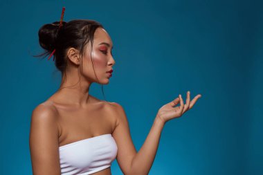 Serious elegant asian girl posing with hand in air and looking at camera. Profile of beautiful young brunette woman wear tank top. Female beauty. Isolated on blue background. Studio shoot. Copy space