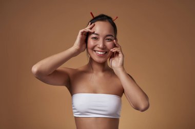 Smiling elegant asian girl touching her face and looking at camera. Beautiful young brunette slim woman wearing tank top. Female beauty. Isolated on orange background. Studio shoot. Copy space