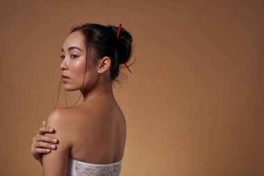 Side view of stylish asian girl hugging herself and looking away. Attractive young brunette woman wearing tank top. Female beauty. Isolated on orange background. Studio shoot. Copy space