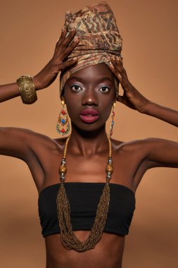 Partial of stylish black girl wear traditional african outfit and accessories look at camera. Young woman wearing tank top, turban, necklace, earrings and bracelet. Orange background. Studio shoot