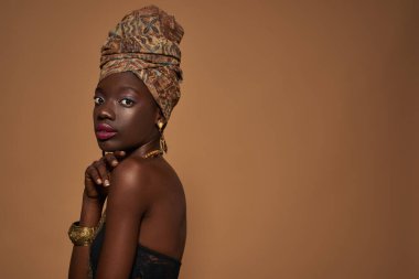 Side view of serious elegant black girl wearing traditional african outfit and accessories looking at camera. Beautiful young slim woman. Isolated on orange background. Studio shoot. Copy space