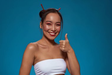 Smiling elegant asian girl showing thumb up gesture and looking at camera. Beautiful young brunette slim woman wearing tank top. Female beauty. Isolated on blue background. Studio shoot. Copy space