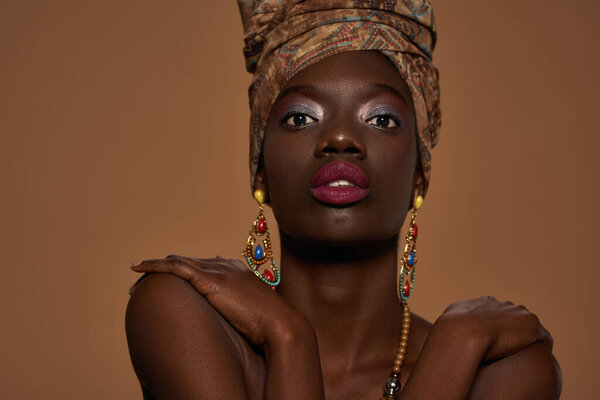 Cropped fashionable black girl wearing traditional african outfit and accessories looking at camera. Pretty young woman wear turban, necklace and earrings. Orange background. Studio shoot. Copy space