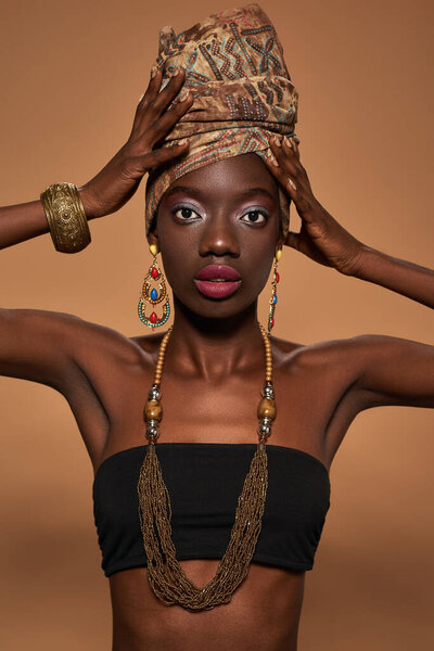Partial of stylish black girl wear traditional african outfit and accessories look at camera. Young woman wearing tank top, turban, necklace, earrings and bracelet. Orange background. Studio shoot