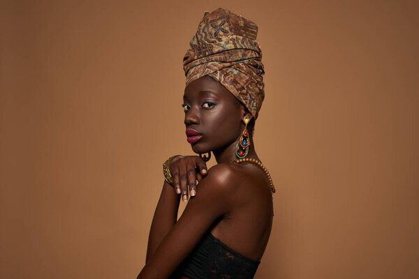Side view of fashionable black girl wearing traditional african outfit and accessories looking at camera. Pretty young slim woman. Isolated on orange background. Studio shoot. Copy space