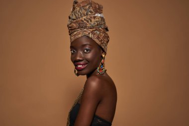 Side view of smiling stylish black girl wearing traditional african outfit and accessories looking at camera. Attractive young slim woman. Isolated on orange background. Studio shoot. Copy space