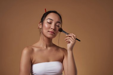 Elegant asian girl with closed eyes applying cosmetic brush on her face. Beautiful young brunette slim woman wearing tank top. Female beauty. Isolated on orange background. Studio shoot. Copy space