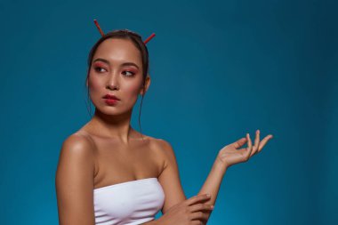 Focused stylish asian girl with hand in air looking away. Attractive young brunette slim woman wearing tank top. Female beauty. Isolated on blue background. Studio shoot. Copy space