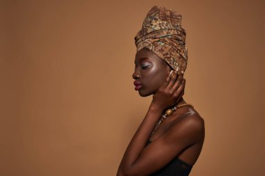 Side view of thoughtful elegant black girl wearing traditional african outfit and accessories. Profile of beautiful young slim woman. Isolated on orange background. Studio shoot. Copy space