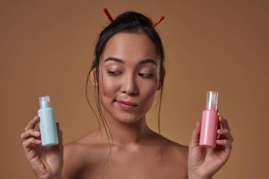 Focused elegant asian girl holding and looking at colorful nail polishes. Beautiful young brunette slim woman. Female beauty. Isolated on orange background. Studio shoot. Copy space