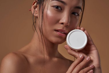 Partial of elegant asian girl holding container with cosmetic cream and looking at camera. Beautiful young brunette slim woman. Body and skin care. Isolated on orange background. Studio shoot
