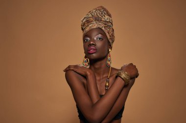 Front view of smiling stylish black girl wearing traditional african outfit and accessories hugging herself and looking at camera. Attractive young slim woman. Orange background in studio. Copy space