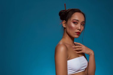 Side view of serious fashionable asian girl looking at camera. Pretty young brunette slim woman wearing tank top. Female beauty. Isolated on blue background. Studio shoot. Copy space