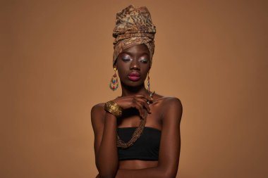 Front view of thoughtful elegant black girl wearing traditional african outfit and accessories. Beautiful young slim woman. Isolated on orange background. Studio shoot. Copy space