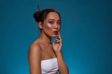 Cropped of serious elegant japanese girl looking at camera. Beautiful young brunette slim woman wearing tank top. Female beauty. Isolated on blue background. Studio shoot. Copy space